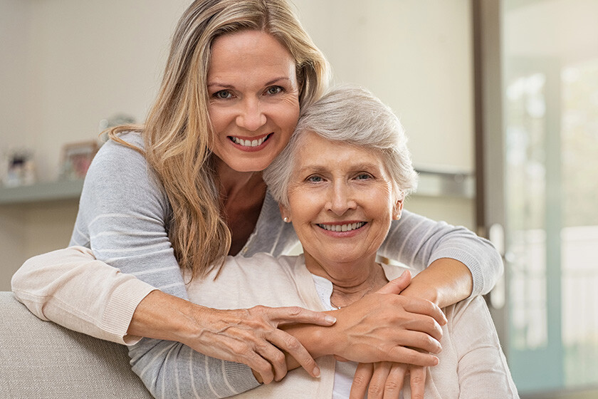 Cheerful mature woman embracing senior mother at home and looking at camera. Portrait of elderly mother and middle aged daughter smiling together. Happy daughter embracing from behind elderly mom sitting on sofa.