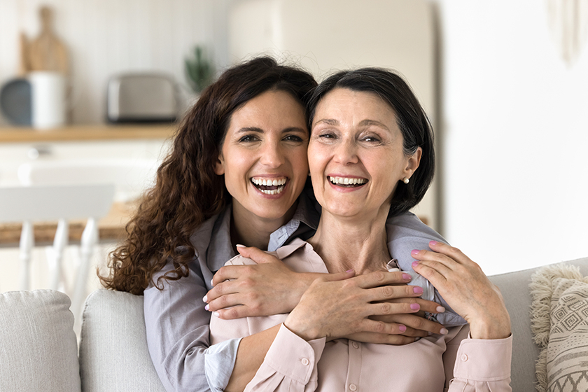 Close up portrait happy different age women hugging at home. Beautiful Hispanic female cuddling tightly her mature mother, laughing, looking at camera enjoy time together, showing strong family ties