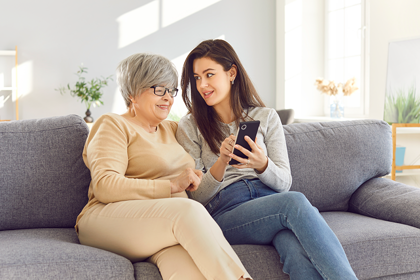 Portrait of an elderly senior woman sitting on sofa in living room at home with her adult daughter and using smartphone together browsing apps. Woman showing her mother video or news on mobile phone. 