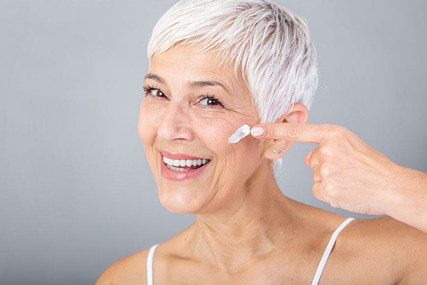 Smiling senior woman applying anti-aging lotion to remove dark circles under eyes. Mature woman using cosmetic cream to hide wrinkles. Lady using day moisturizer to counteract the aging of the skin.