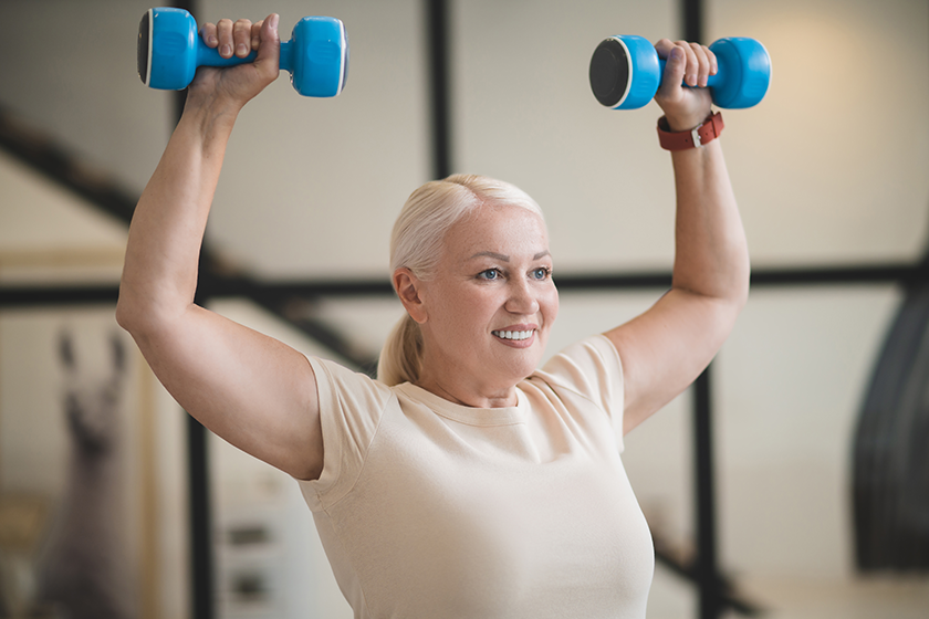 Waist-up portrait of a smiling pleased beautiful mature fit lady performing a dumbbell overhead press 