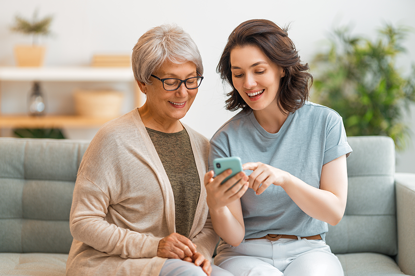 Young and senior women are using smartphone. Daughter is helping mother having fun staying at home.